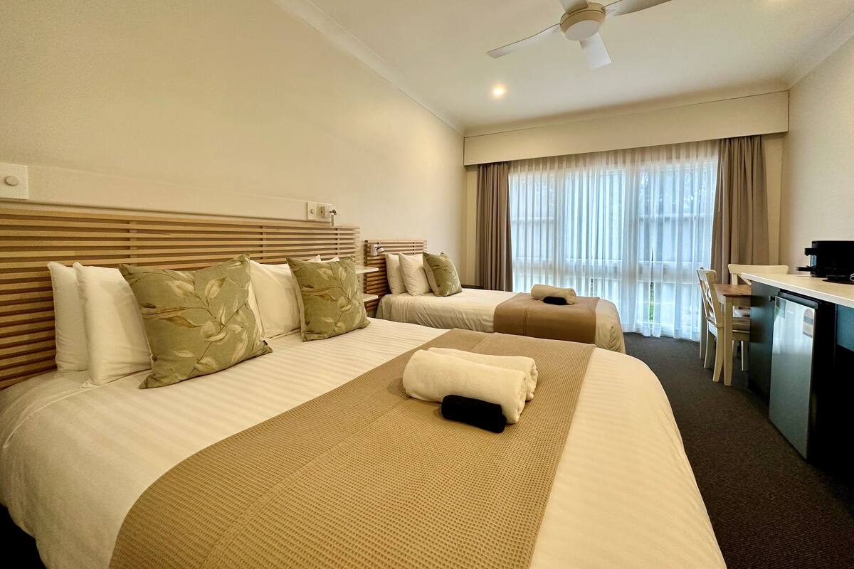 A hotel room with two beds, a desk, a chair, a ceiling fan, and a window with curtains.