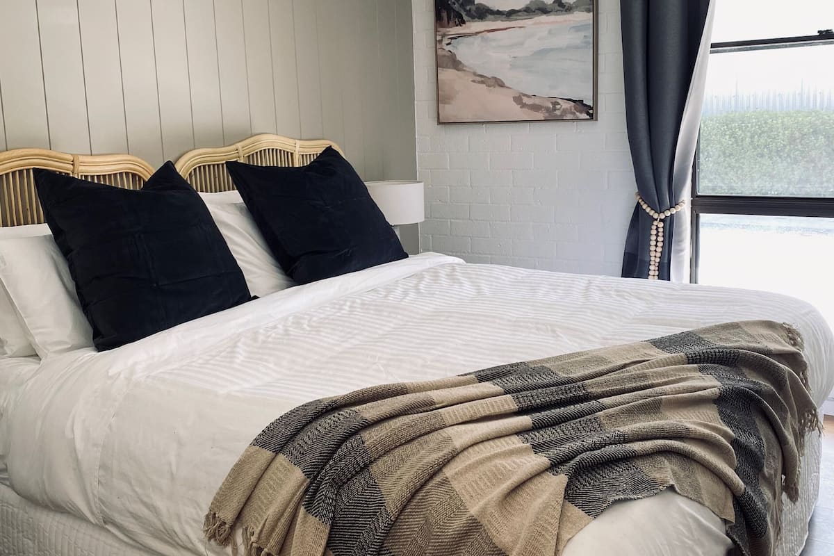 A bedroom with a bed, pillows, a blanket, a painting on the wall, and a window with curtains.