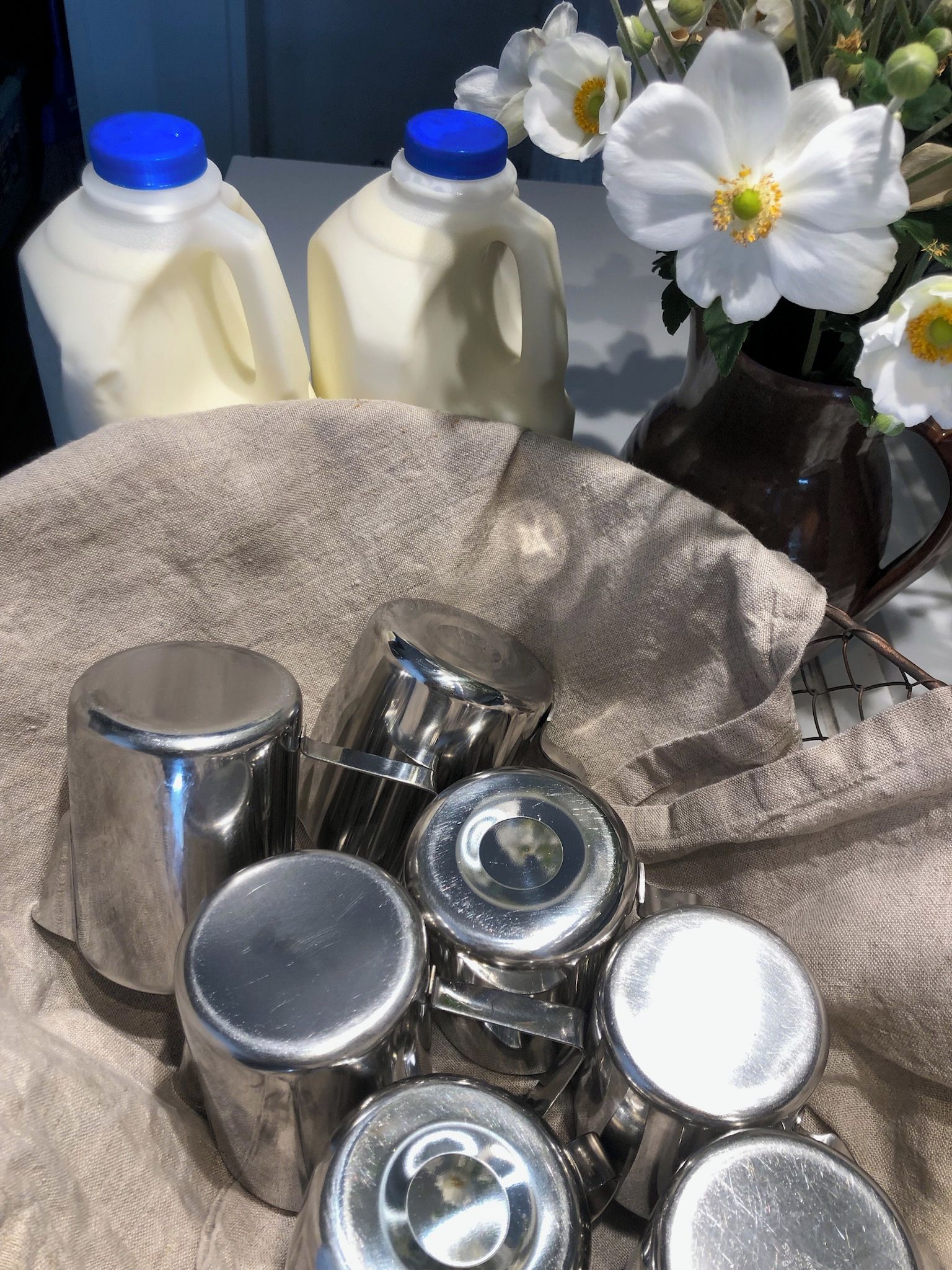 Una mesa con botellas de leche, recipientes metálicos y un florero con flores blancas.