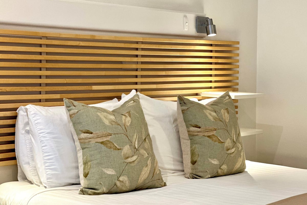 A bed with wooden slats headboard, white and patterned pillows, and a bedside table with a lamp.