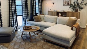 A modern living room with a sectional sofa, a small round coffee table, and a grey armchair. There is a wooden cabinet with decorative items and a plant on the wall. - Berrima Bakehouse Motel (Berrima)