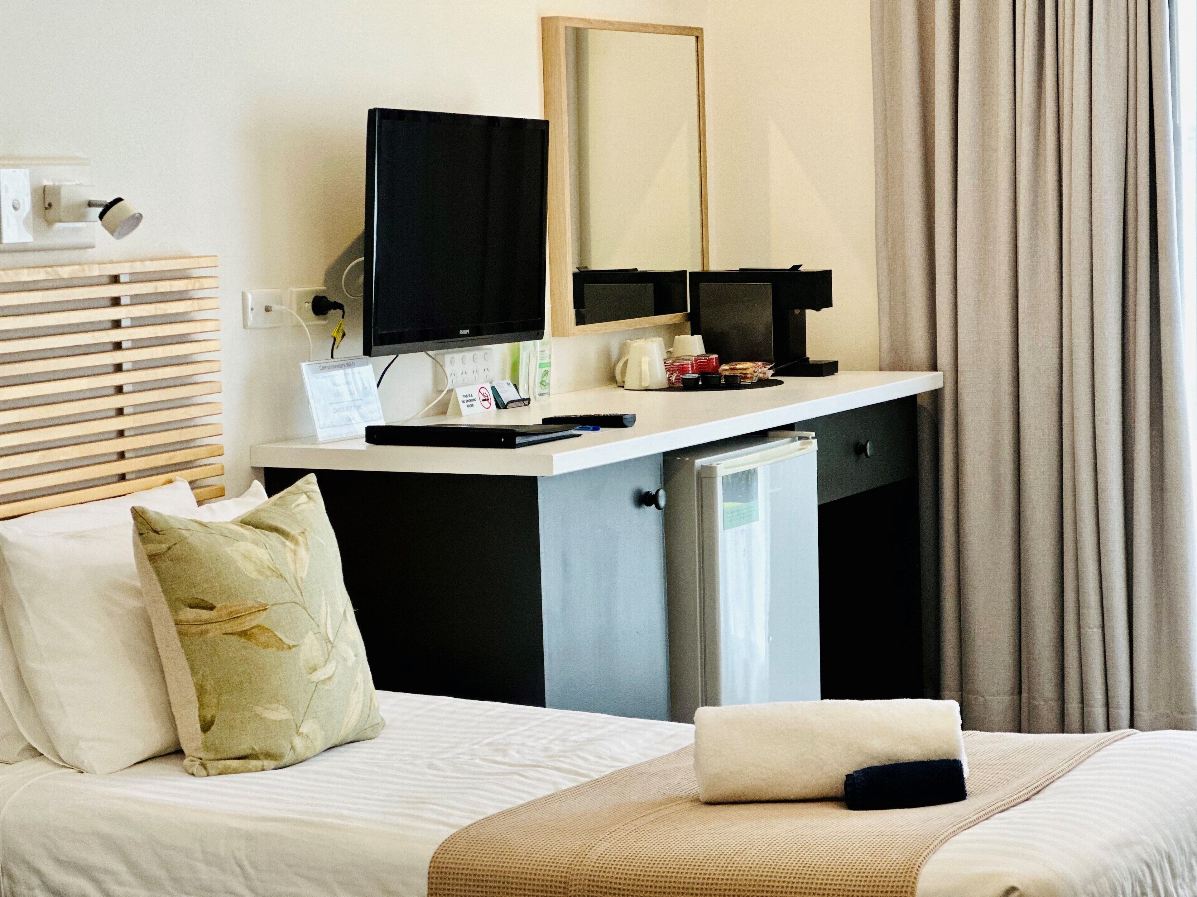 A hotel room with a bed, a desk with a computer, a small fridge, a mirror, and a window with curtains.