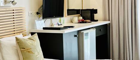 A hotel room with a bed, a desk with a computer, a small fridge, a mirror, and a window with curtains.