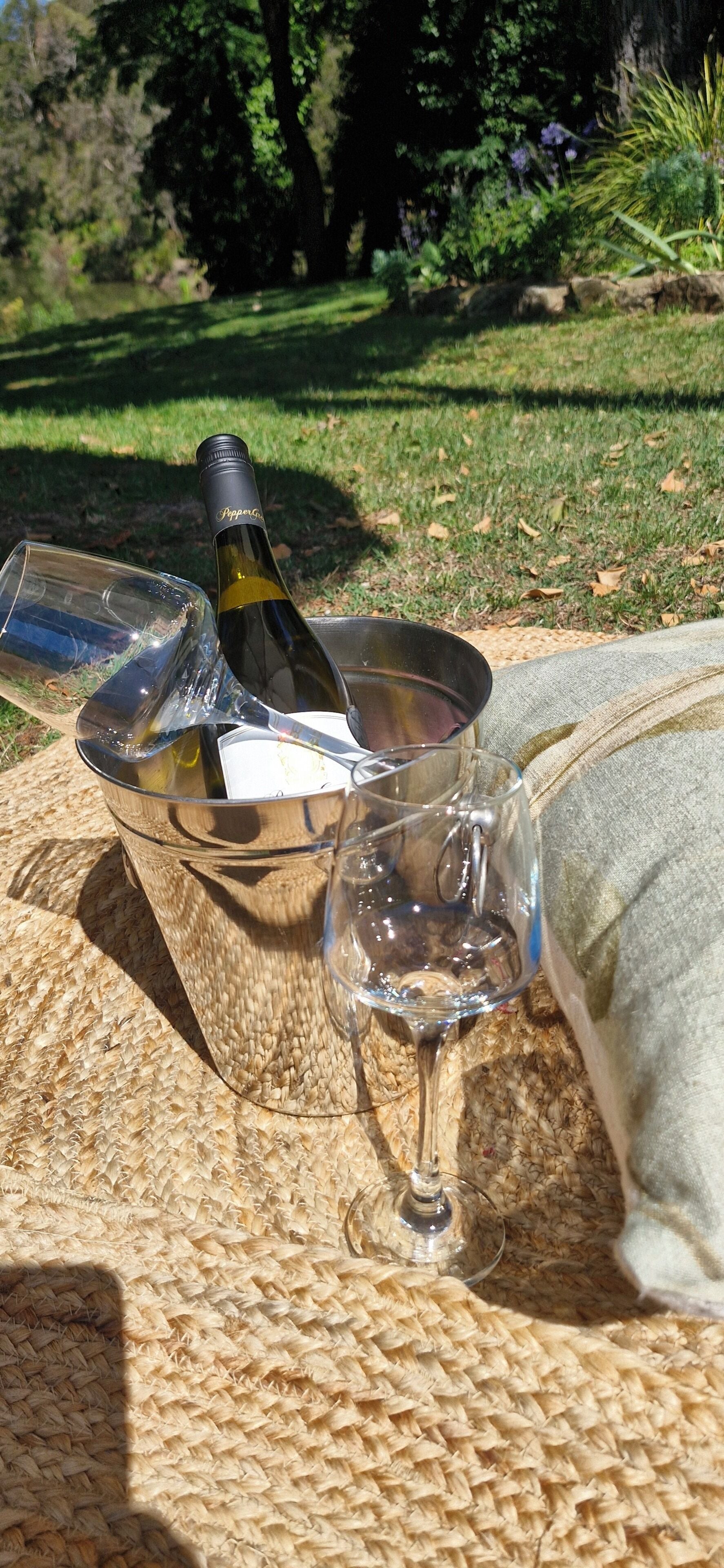 A bottle of wine, a glass, and a wine glass on a woven mat outdoors.
