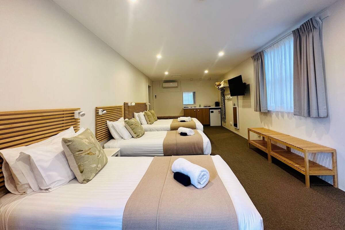 A hotel room with two beds, a small kitchenette, a TV, and a window with curtains.