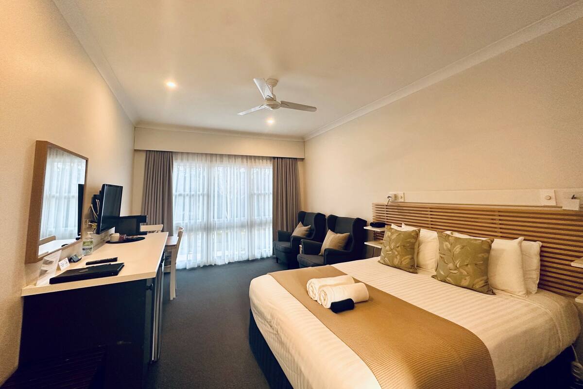 A hotel room with a bed, a desk, a chair, a ceiling fan, and a window with curtains.