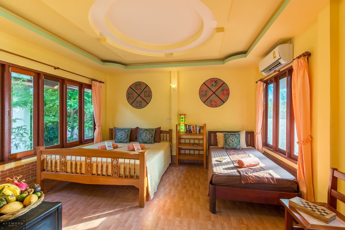A room with two beds, a small table, a shelf with books, and a window with curtains.