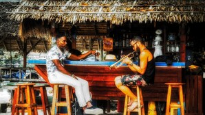 Two people sitting at a bar with a thatched roof, wooden stools, and a shelf with bottles. - Seaboard Bungalows (Ko Pha-ngan)