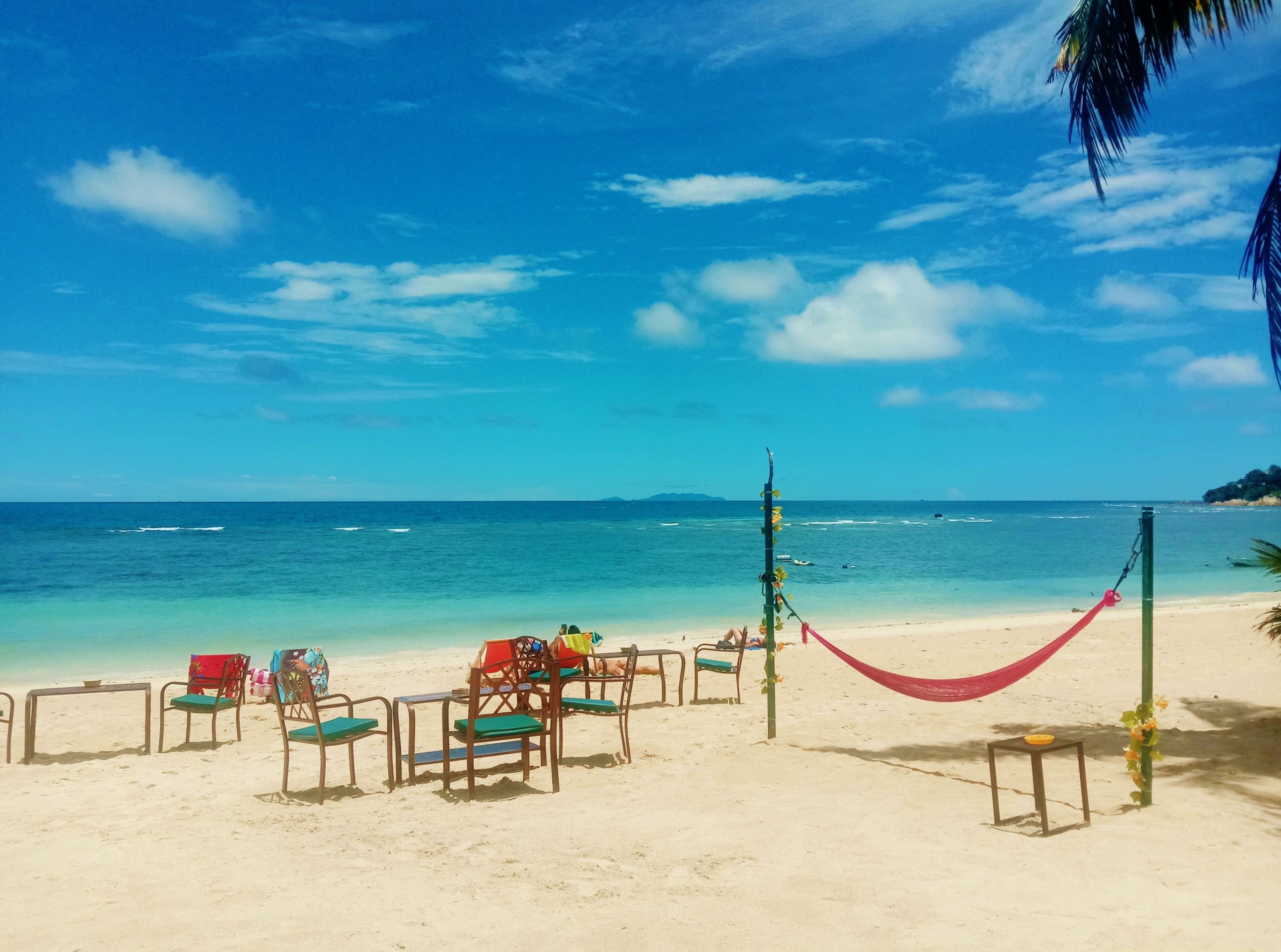 A beach with chairs and tables, a hammock, and a pole with flowers.