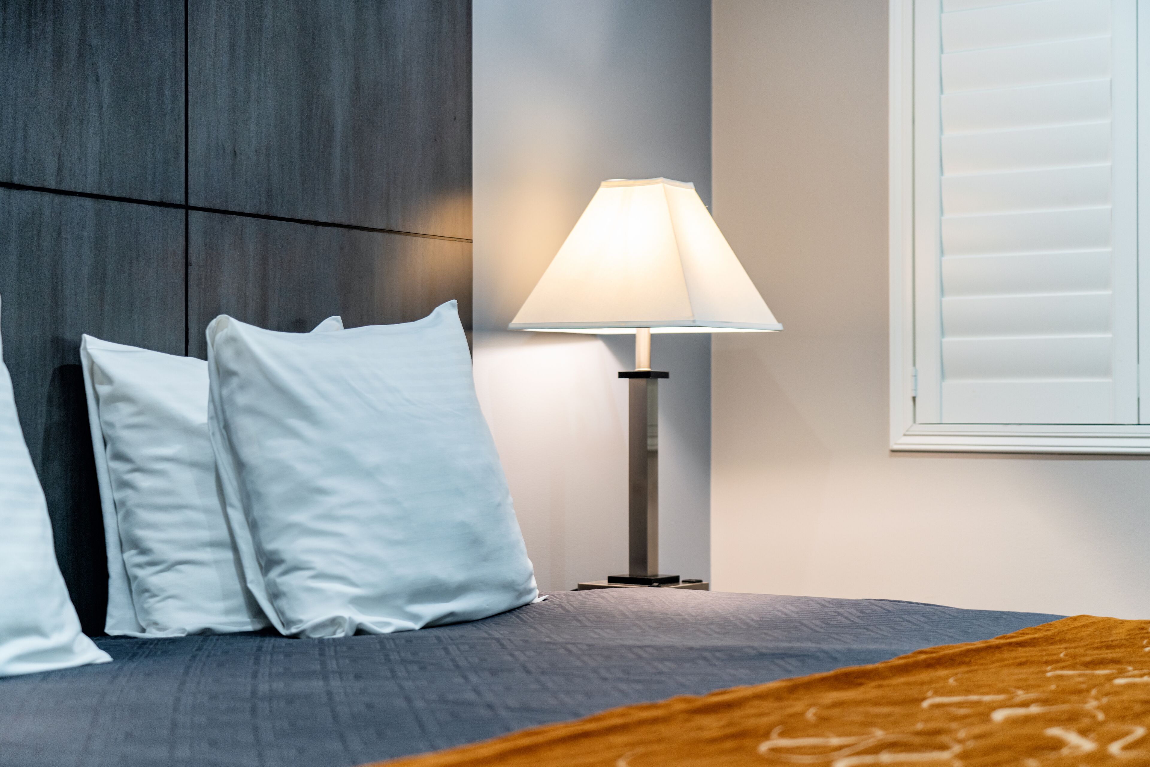 A neatly made bed with white pillows, a grey headboard, a floor lamp, and a window with blinds.