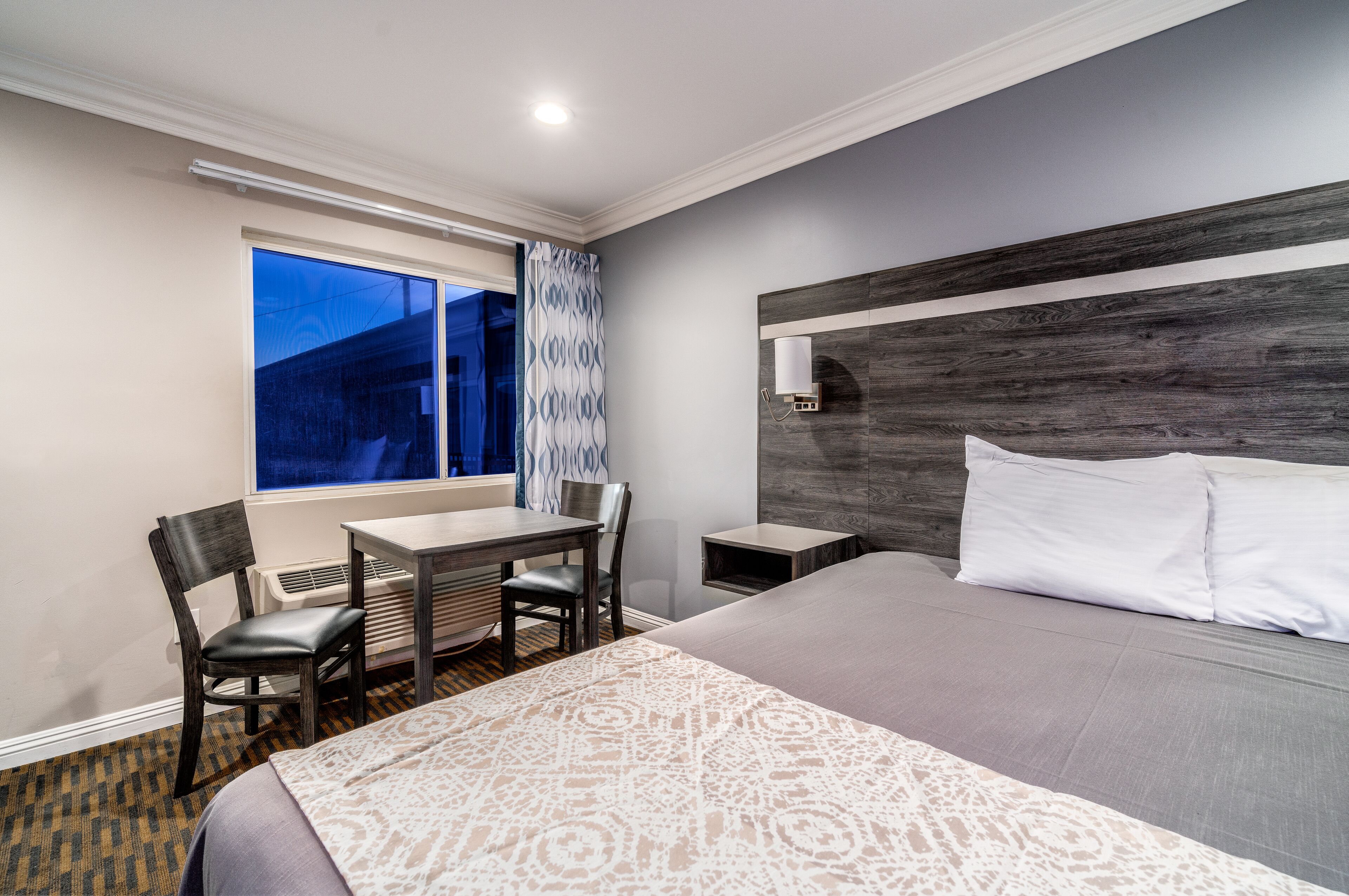 A hotel room with a large bed, a desk with two chairs, a window with a view, and a wall-mounted nightstand.
