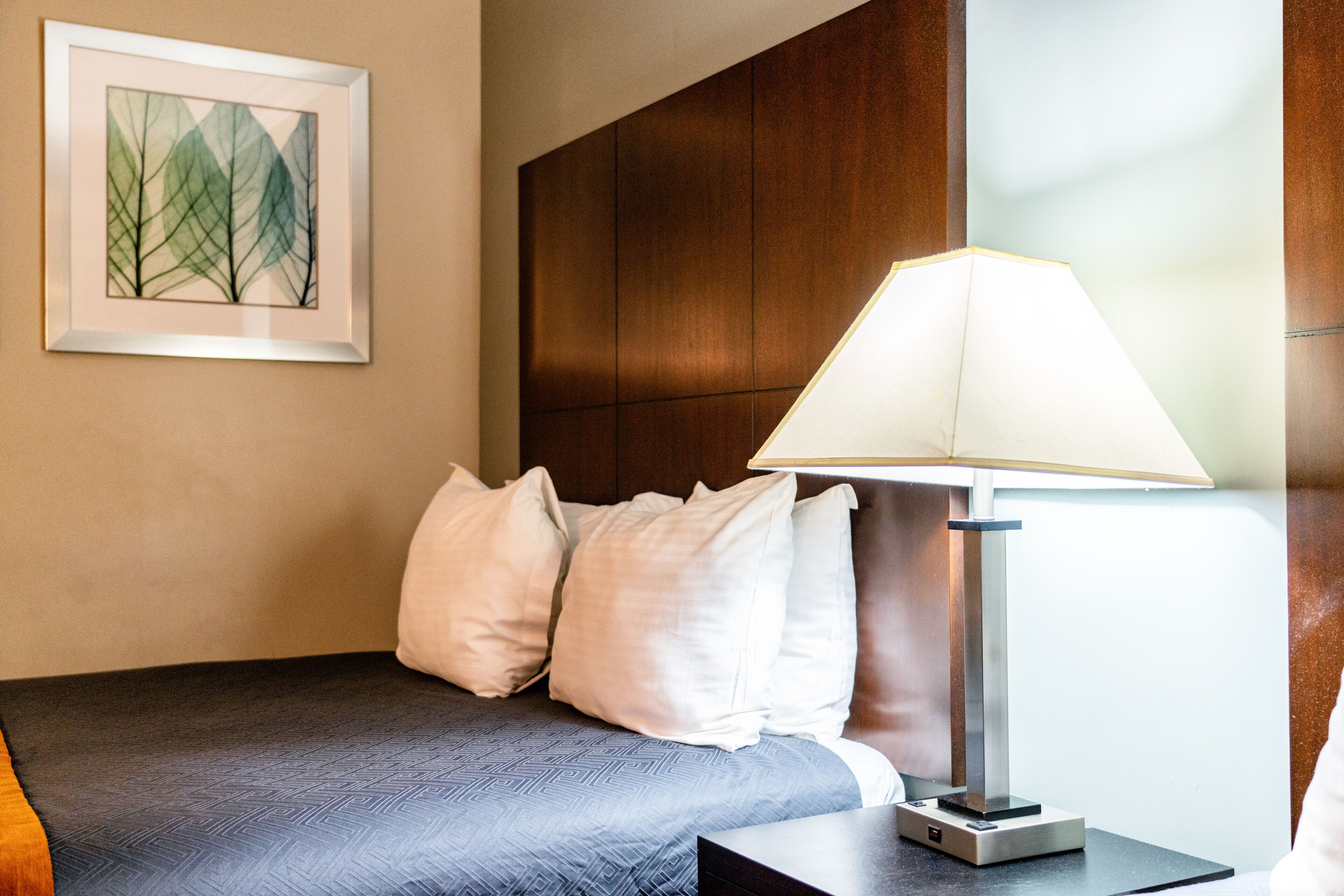 A hotel room with a bed, a bedside lamp, a framed artwork on the wall, and a wooden headboard.