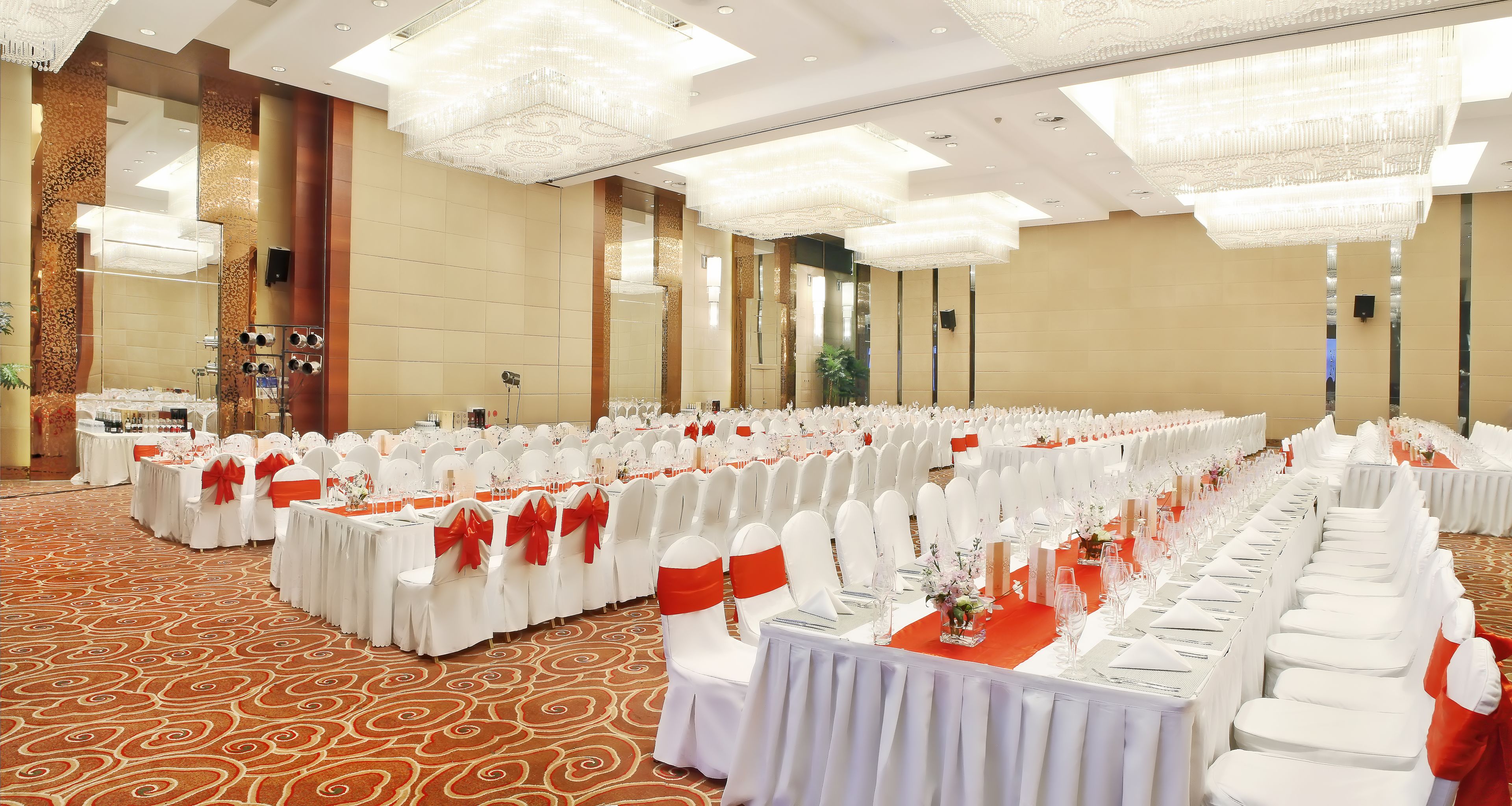 Un salón de banquetes con filas de mesas, sillas blancas con cintas rojas y una alfombra estampada.