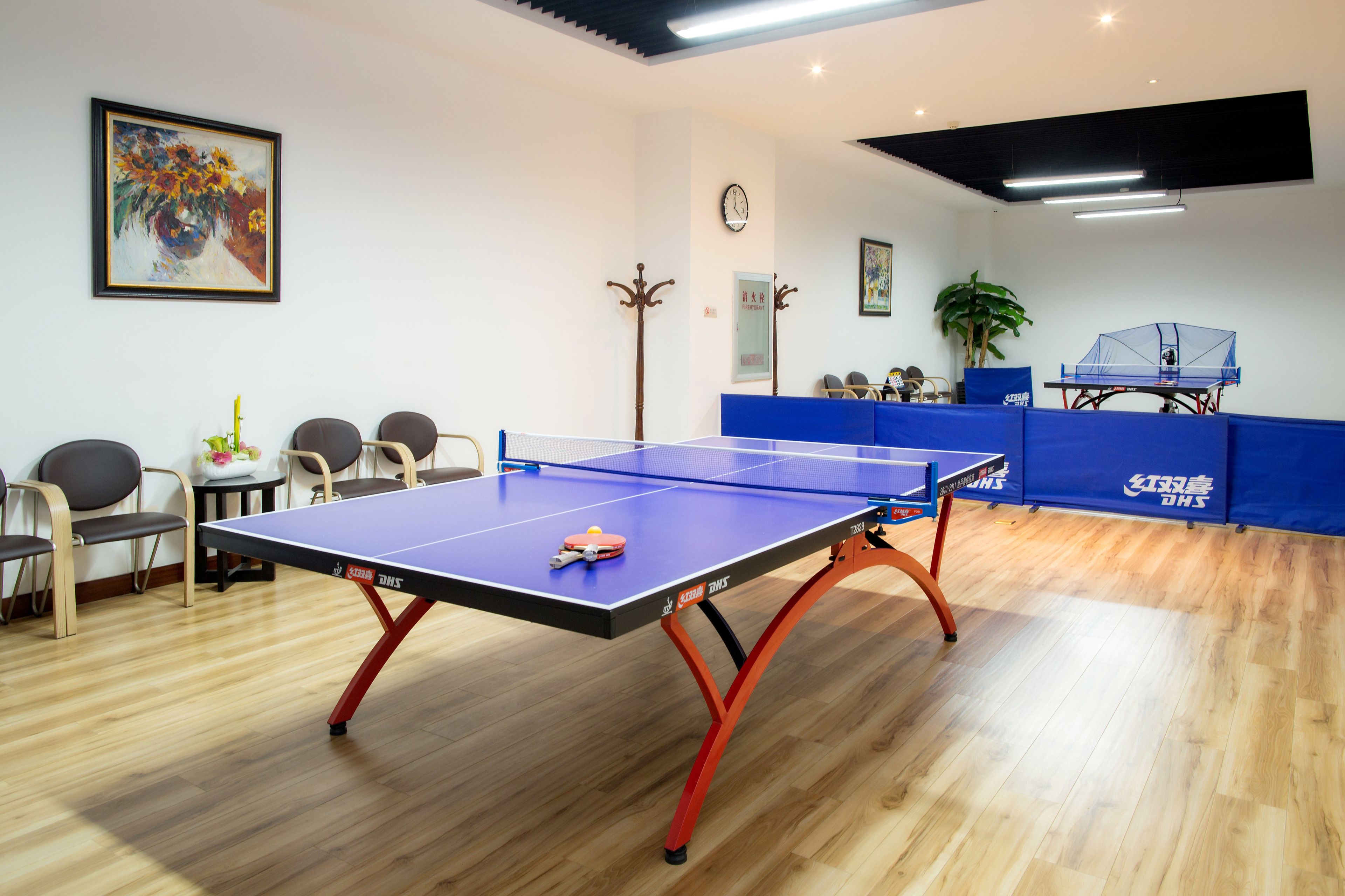 A ping pong table in a room with wooden flooring, chairs, and a clock on the wall.
