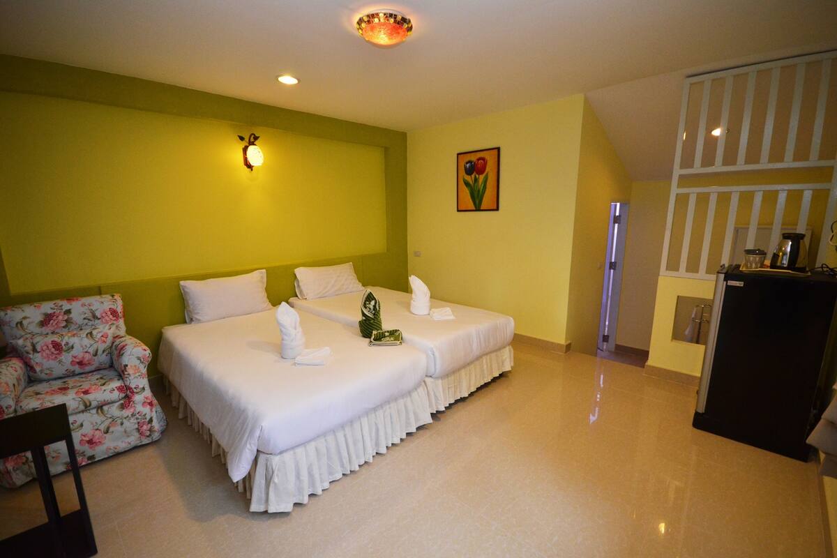 A hotel room with two beds, a floral armchair, a painting of flowers, and a kitchenette.