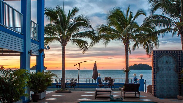 Zwembadterras met uitzicht op de oceaan bij zonsondergang, palmbomen en een gebouw met blauw-witte gevel.