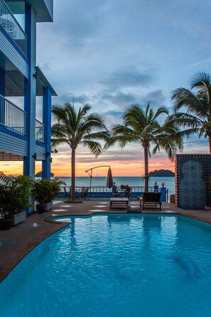 Un espace aménagé au bord de la piscine, offrant une vue sur l’océan au coucher du soleil, avec des palmiers et un bâtiment à la façade bleue et blanche.