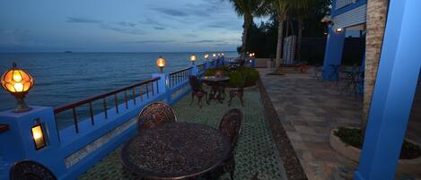 An outdoor dining area with a view of the ocean, featuring a blue railing, metal tables, and chairs.