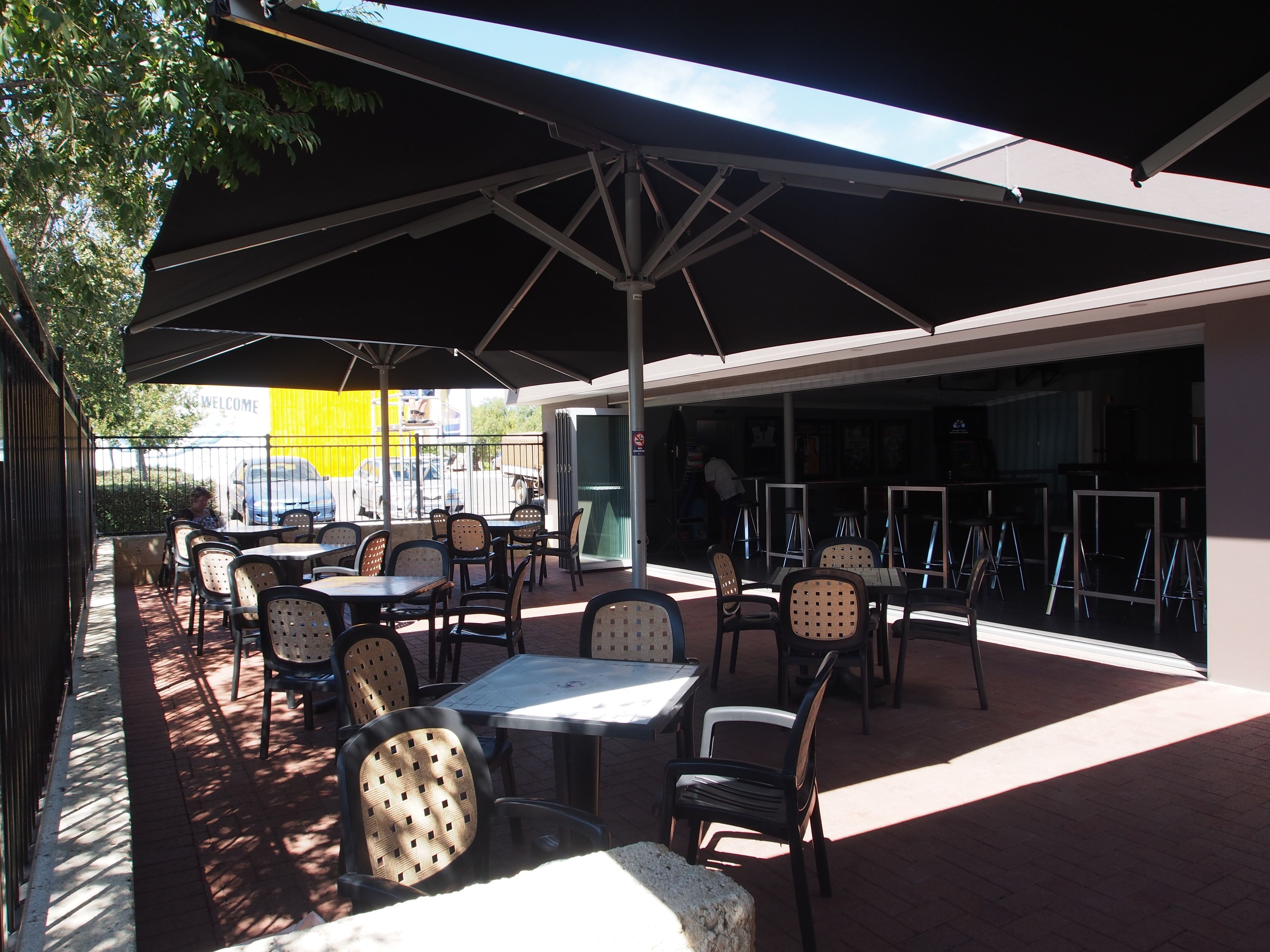 Une terrasse extérieure avec des tables et des chaises disposées sous un grand parasol, entourée d’un sol en briques et d’une clôture en métal.