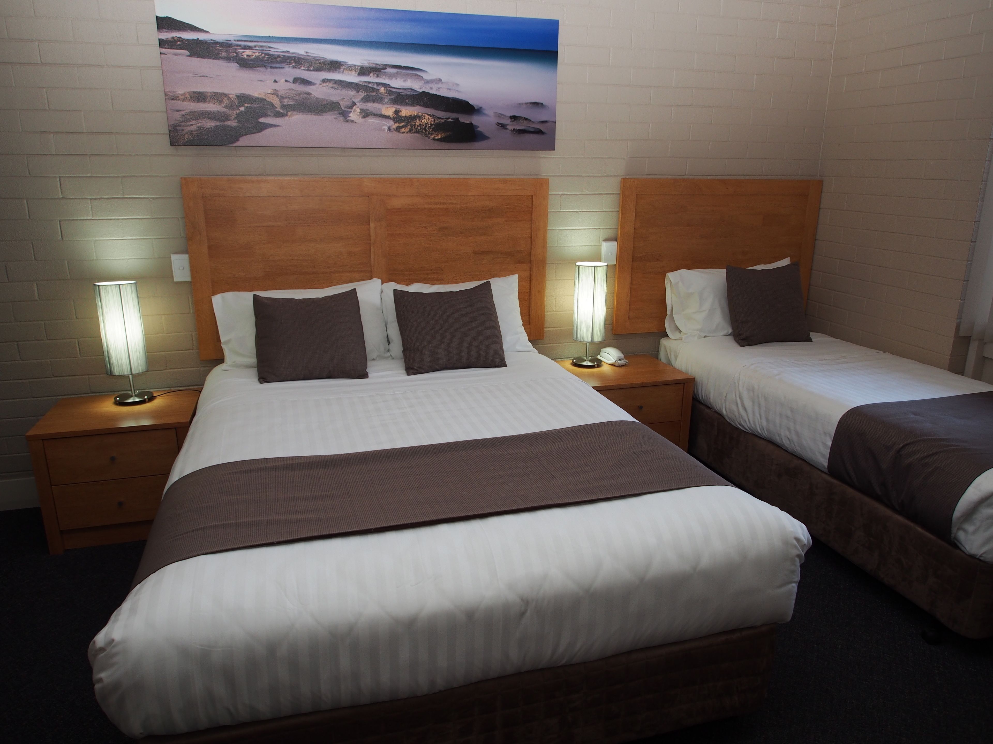 A hotel room with two beds, each with a wooden headboard, white bedding, and a nightstand with a lamp. A painting of a beach scene is on the wall above the beds.