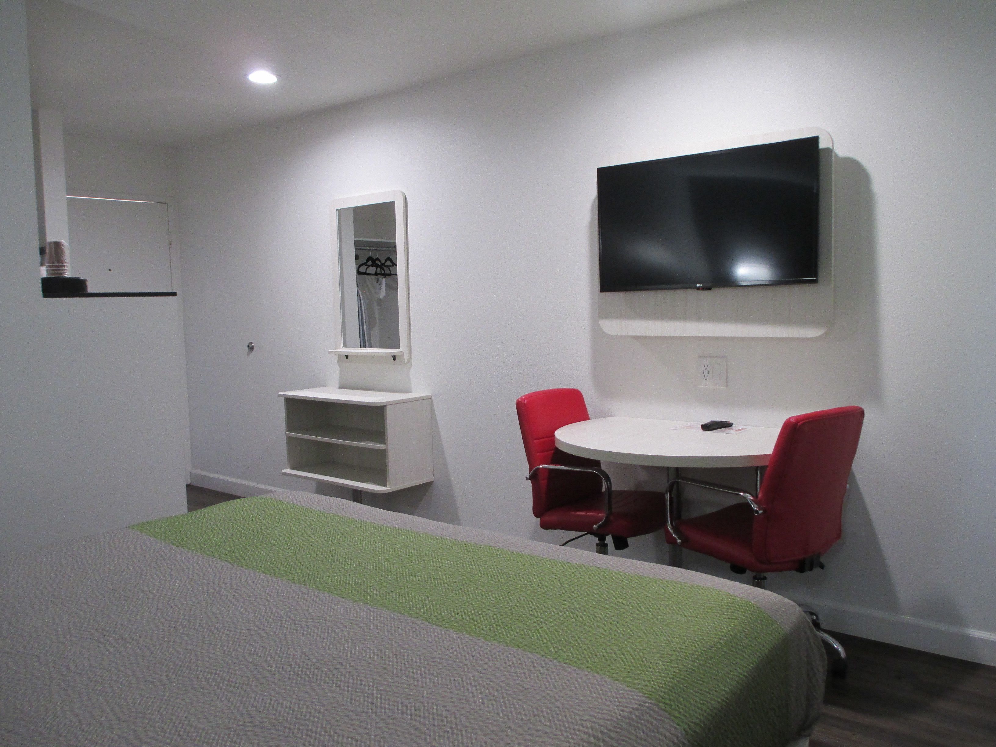 A hotel room with a bed, a small desk with two red chairs, a TV mounted on the wall, and a mirror above the desk.