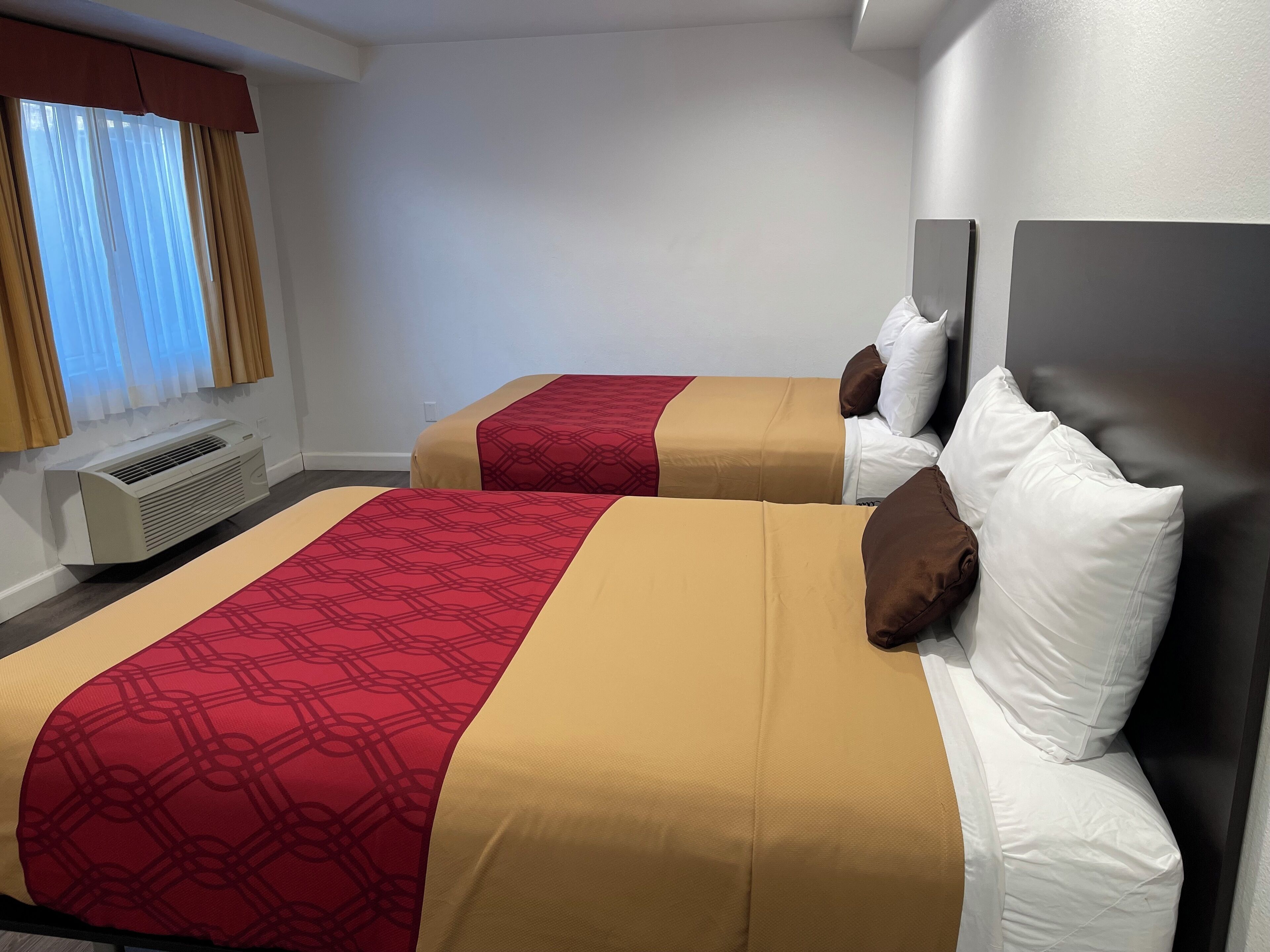 A hotel room with two beds, each with a red and beige bedspread, white pillows, and a wall-mounted air conditioner.
