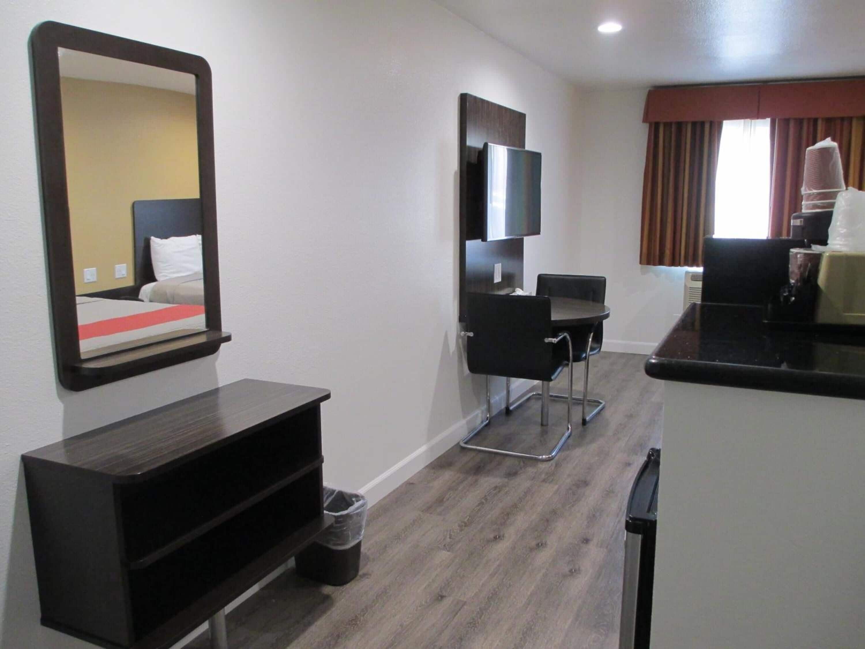 A hotel room with a bed, a TV mounted on the wall, a small desk with a chair, a trash bin, and a kitchenette area.