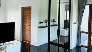 A modern living room with a flat-screen TV, a sofa, two wooden tables, a glass partition, and a bathroom visible through the partition. - Ice Kamala Beach Hotel (Kamala)