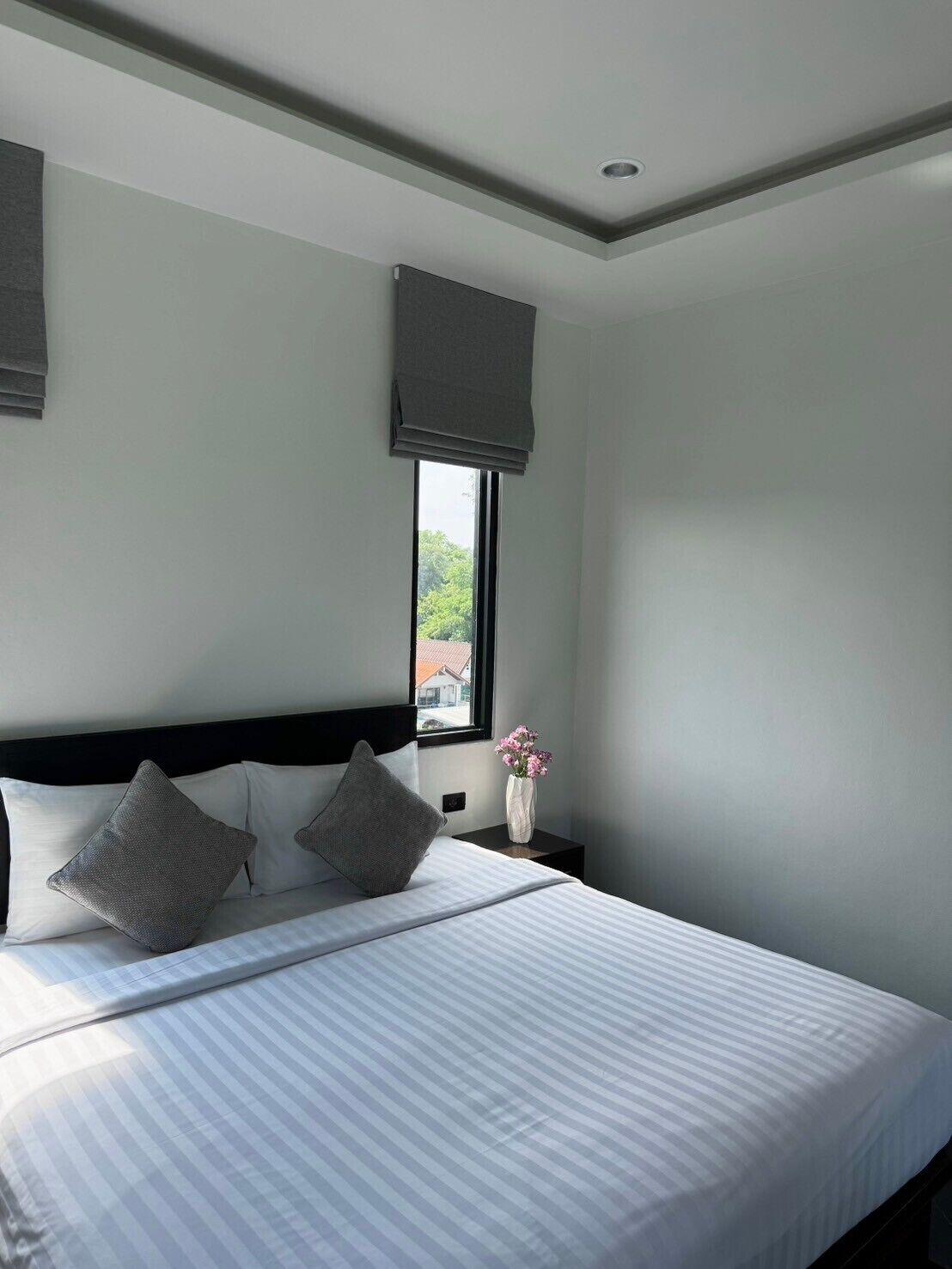 A neatly made bed with striped bedding, a dark headboard, and two pillows. A window with a view of greenery and buildings is visible, and a vase with flowers is on the nightstand.