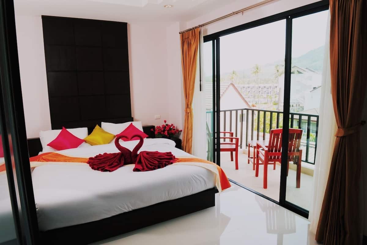 Une chambre d’hôtel avec un grand lit, un balcon avec des chaises rouges et une vue sur des immeubles et des collines.