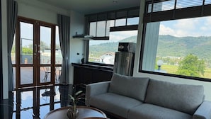 A modern living room with a sofa, two round tables, a view of a mountainous landscape through large windows, and a kitchen area with a refrigerator. - Ice Kamala Beach Hotel (Kamala)