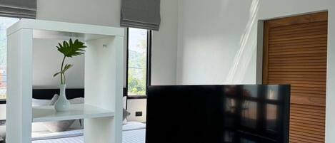 Un soggiorno moderno con una televisione a schermo piatto su un mobile bianco, una libreria con libri, una pianta in vaso e una finestra vista verde.