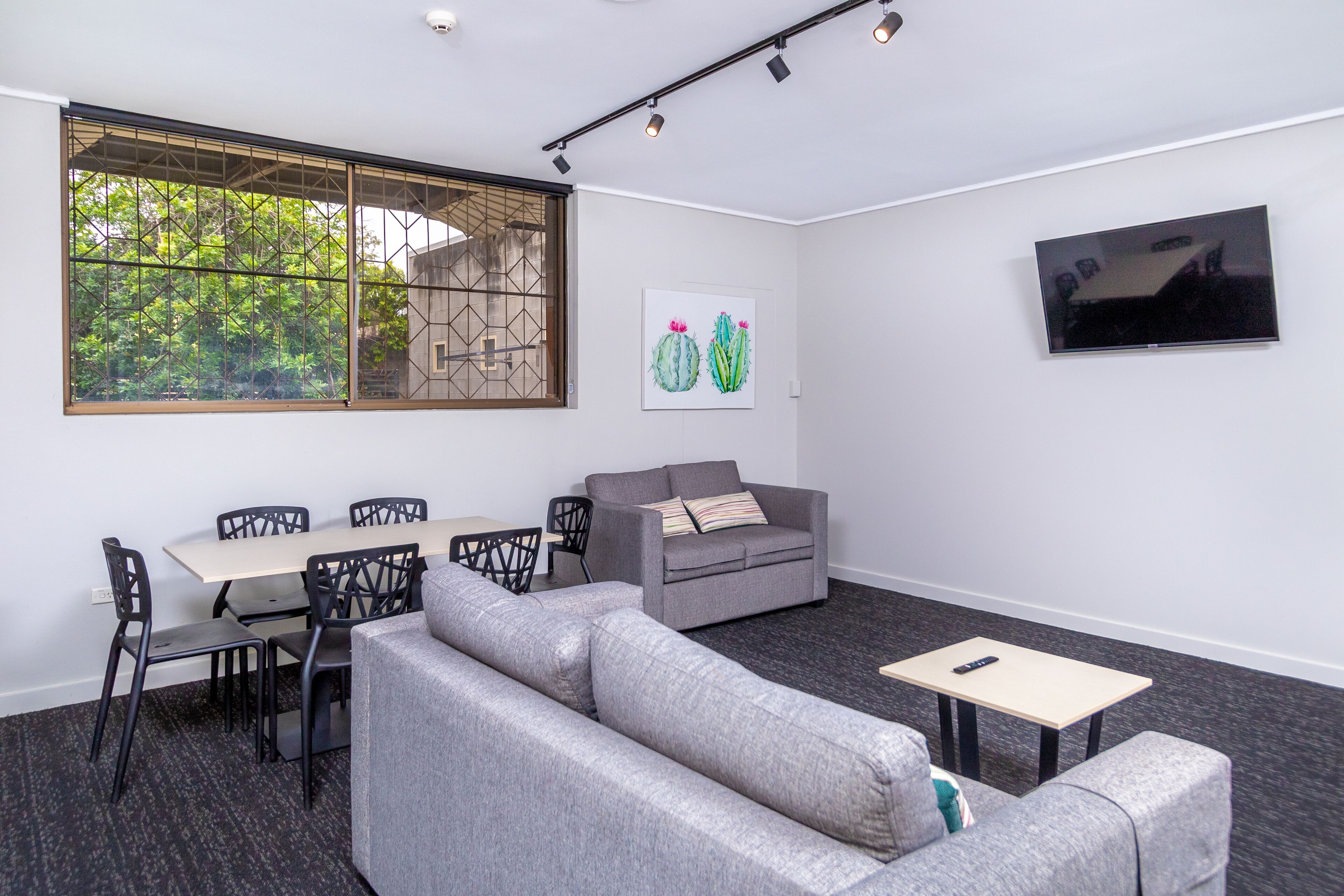 Una habitación moderna con sofá, una mesa de comedor con sillas, televisor montado en la pared y un ventanal con vistas a árboles.