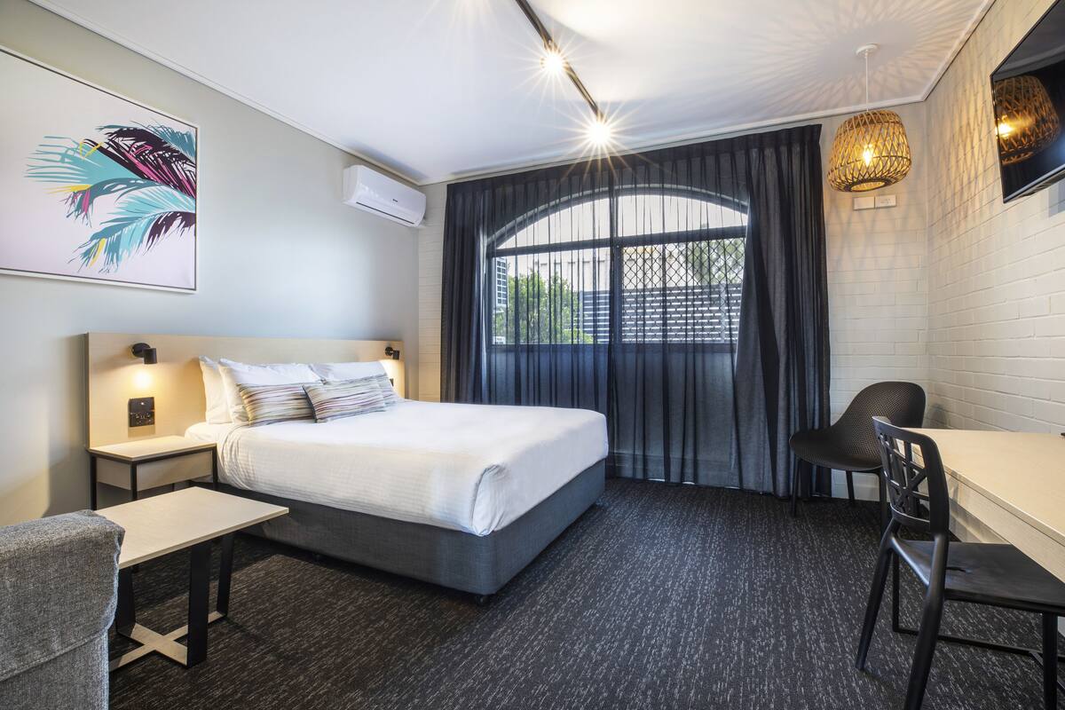 A hotel room with a bed, a desk, a chair, a window with curtains, and a wall-mounted air conditioner.