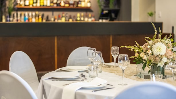 A restaurant setting with a bar in the background, tables set with white tablecloths, and a vase of flowers on the table.