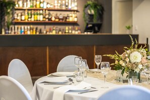 Une salle de restaurant avec un bar en arrière-plan, des tables recouvertes de nappes blanches et un vase de fleurs posé sur chaque table.
