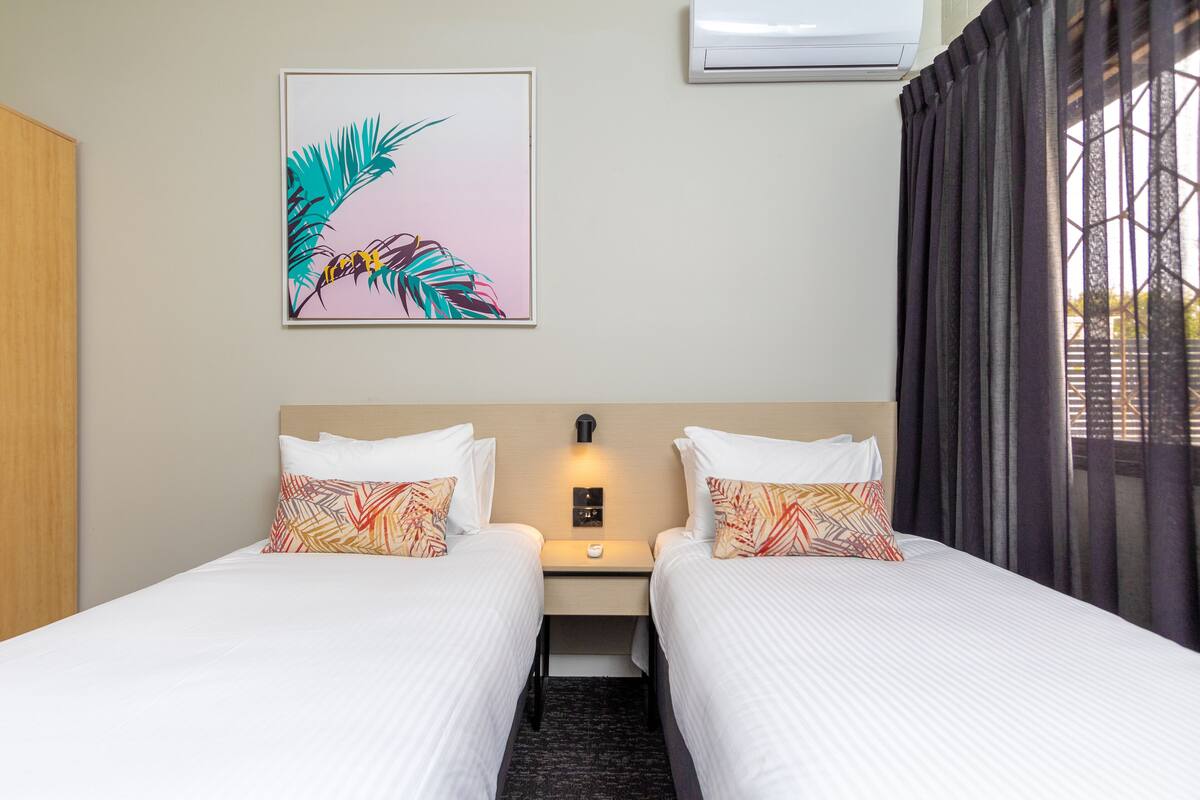 A hotel room with two beds, a nightstand, a wall-mounted air conditioner, and a window with curtains.