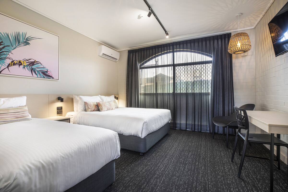 A hotel room with two beds, a desk, a chair, a TV, and a large window with curtains.