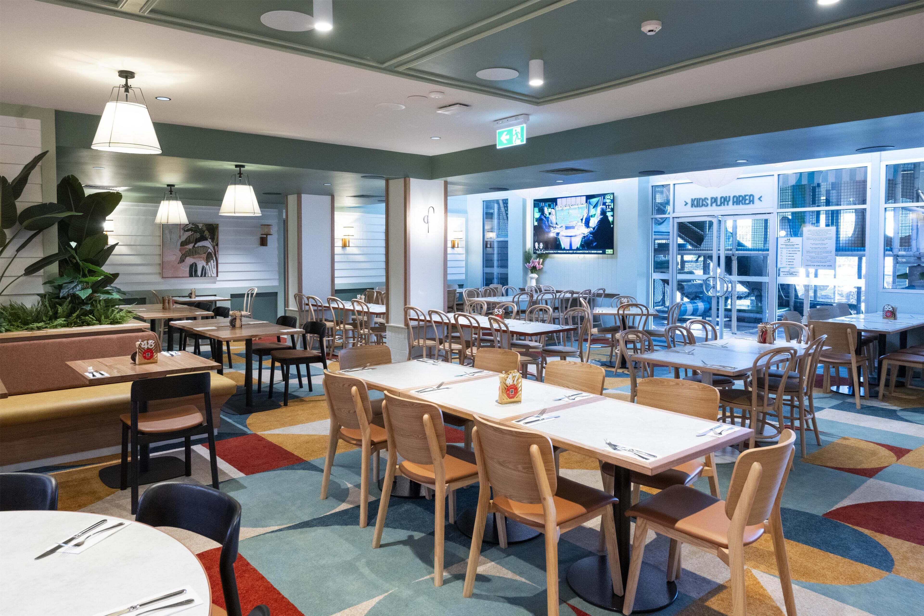 A restaurant with multiple dining tables and chairs, a children's play area, and a television screen displaying a program.