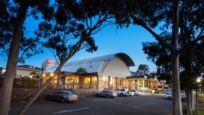 A modern building with a curved roof, surrounded by trees and a parking lot with several cars.