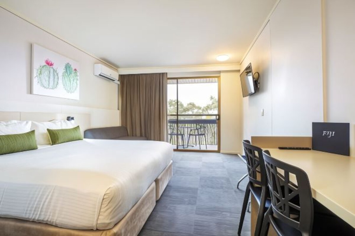 Ein Hotelzimmer mit einem Bett, einem Schreibtisch, einem Stuhl, einem Balkon mit Tisch und Stühlen sowie einem Bild von Kakteen an der Wand.