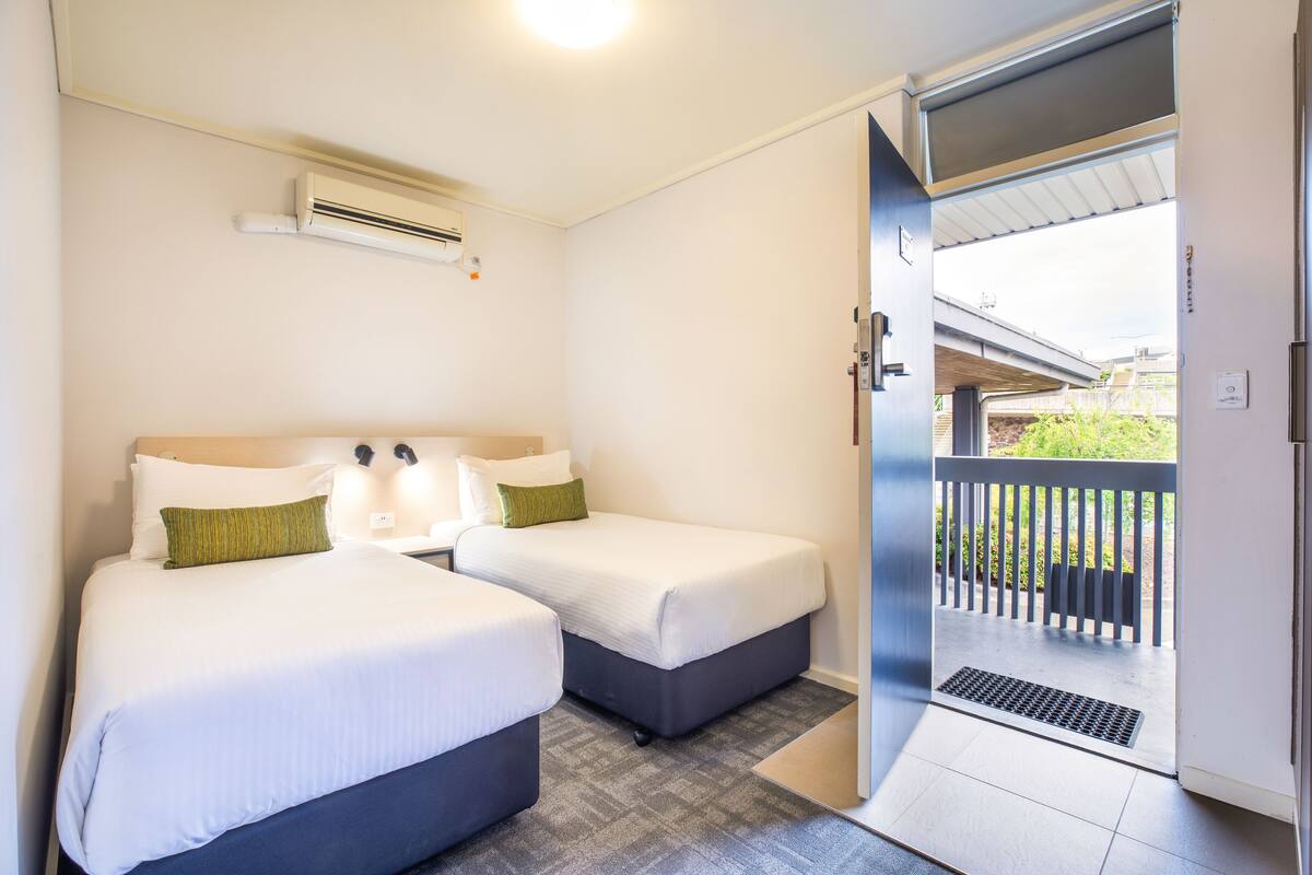 A hotel room with two beds, a ceiling fan, a window with a view of a balcony, and a door leading outside.