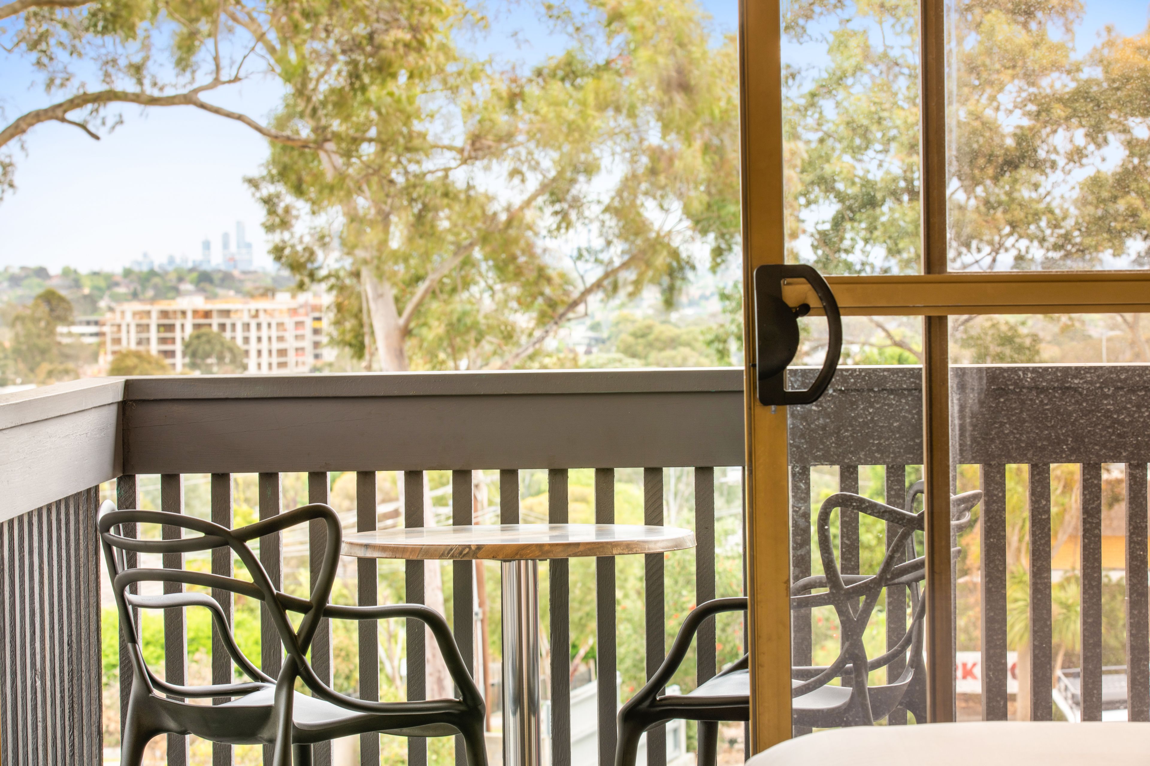 A balcony with a table and chairs, a view of trees and buildings, and a door with a handle.