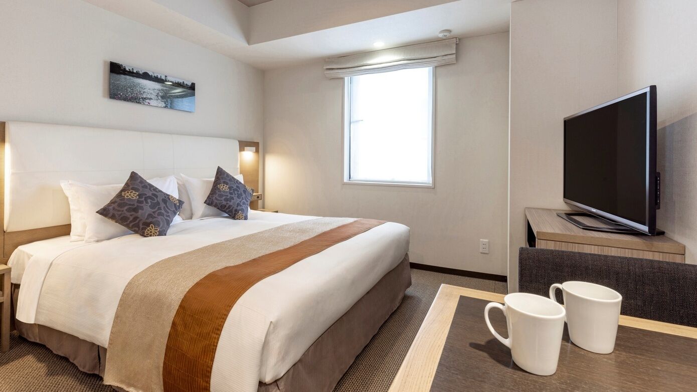 A hotel room with a bed, a TV, a table with two mugs, and a window.