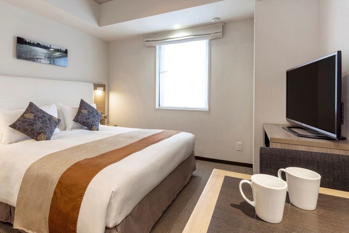 Quarto de hotel com uma cama, uma televisão, uma mesa com duas canecas e uma janela.