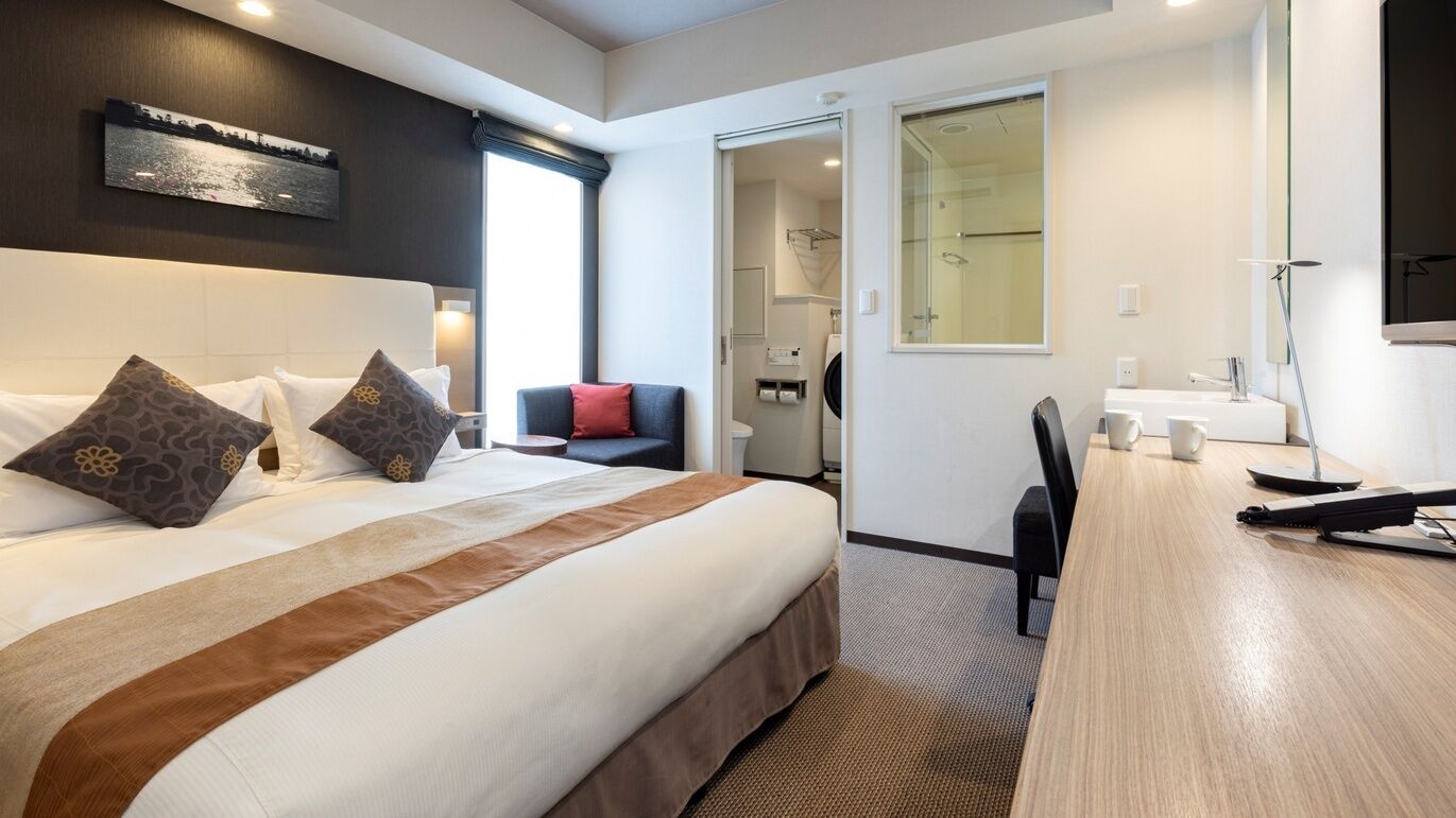 A hotel room with a large bed, a desk, a chair, a small table, a mirror, and a window with a view.