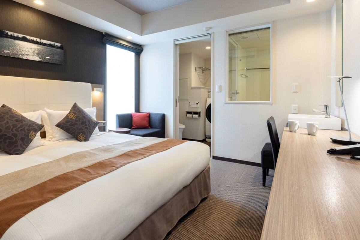 Quarto de hotel com uma cama grande, uma mesa, uma cadeira, uma pequena mesa, um espelho e uma janela com vista.