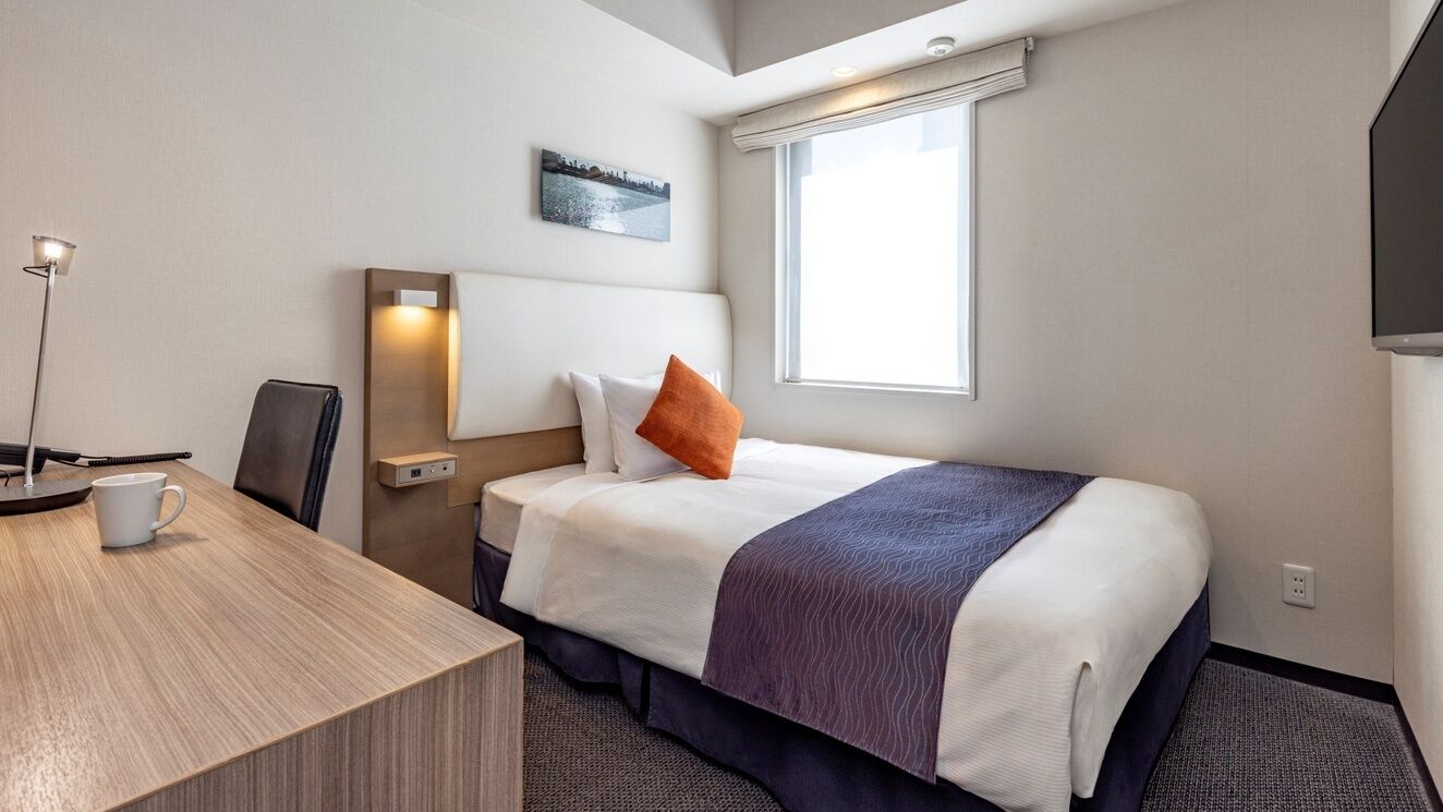 A hotel room with a bed, a desk, a chair, a lamp, a window, and a flat-screen TV.