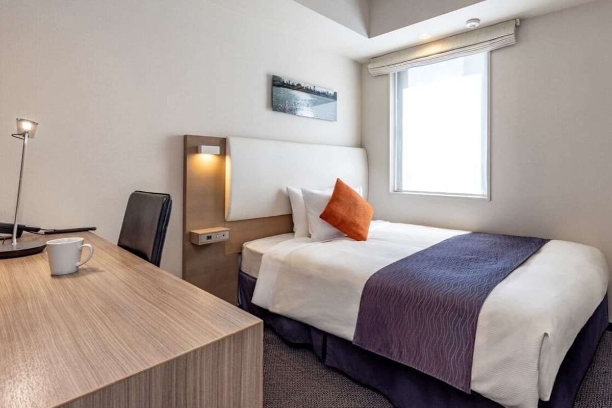 Quarto de hotel com cama, escrivaninha, cadeira, abajur, janela e televisão de tela plana.