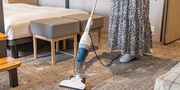 A hotel room with a bed, two ottomans, a standing vacuum cleaner, and a wooden bench.